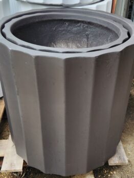 The GardenLite Grecian Cylinder Black S 39x39cm planter, featuring a dark matt finish and vertical ridges, is shown outdoors on a wooden pallet.