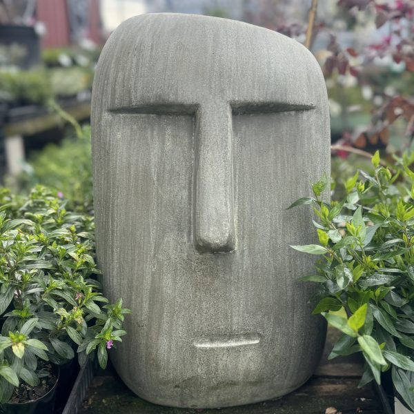 The Moai Statue Green (30x29x46cm), a large, minimalist stone sculpture of a human face reminiscent of the iconic Moai statues, is surrounded by lush green plants in an outdoor setting.