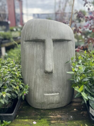 The Moai Statue Green (30x29x46cm), a large, minimalist stone sculpture of a human face reminiscent of the iconic Moai statues, is surrounded by lush green plants in an outdoor setting.