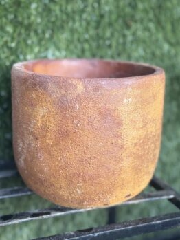 A large, round Portica Egg Pot Rust M 16x13cm with a rough texture sits on a black metal stand against a background of green artificial grass, making it an ideal garden planter.