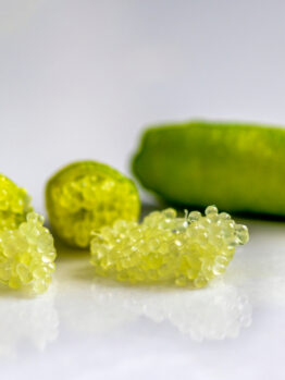 Close-up of finger limes with one cut open, revealing pearl-like citrus vesicles on a smooth white surface.