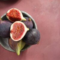 A bowl featuring a mix of whole and halved Ficus 'Violette De Bordeaux' figs sits on a pink surface.