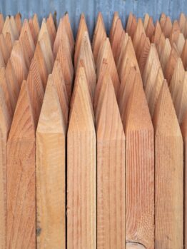 A close-up of Garden Stake Timber (25mmx25mmx1500mm) with pointed tops, stacked vertically before a corrugated metal wall—ideal for garden use.