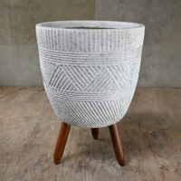 A textured gray concrete planter with geometric patterns stands on three wooden legs against a wooden floor and plain wall background.