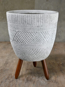 A textured gray concrete planter with geometric patterns stands on three wooden legs against a wooden floor and plain wall background.