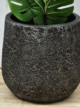 A large, textured dark gray planter with a green monstera plant, placed on a wooden surface against a light-colored wall.