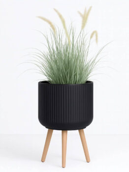 The GardenLite Corrugated Cylinder with Legs Black M 39x38cm planter holds tall ornamental grass with thin green leaves and feathery beige plumes, displayed against a white background.