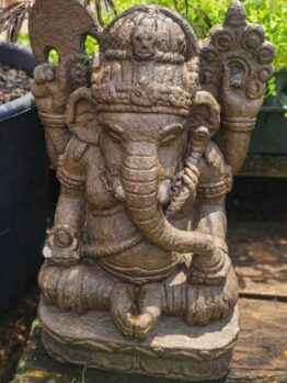 A stone statue of Ganesha, the Hindu deity with an elephant head, seated with one hand raised and detailed carvings, surrounded by greenery.