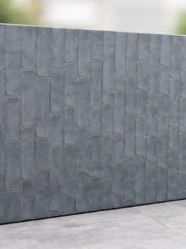 A large rectangular grey concrete wall with a textured, repeating bamboo-like pattern stands outdoors on a paved surface, with blurred greenery in the background.