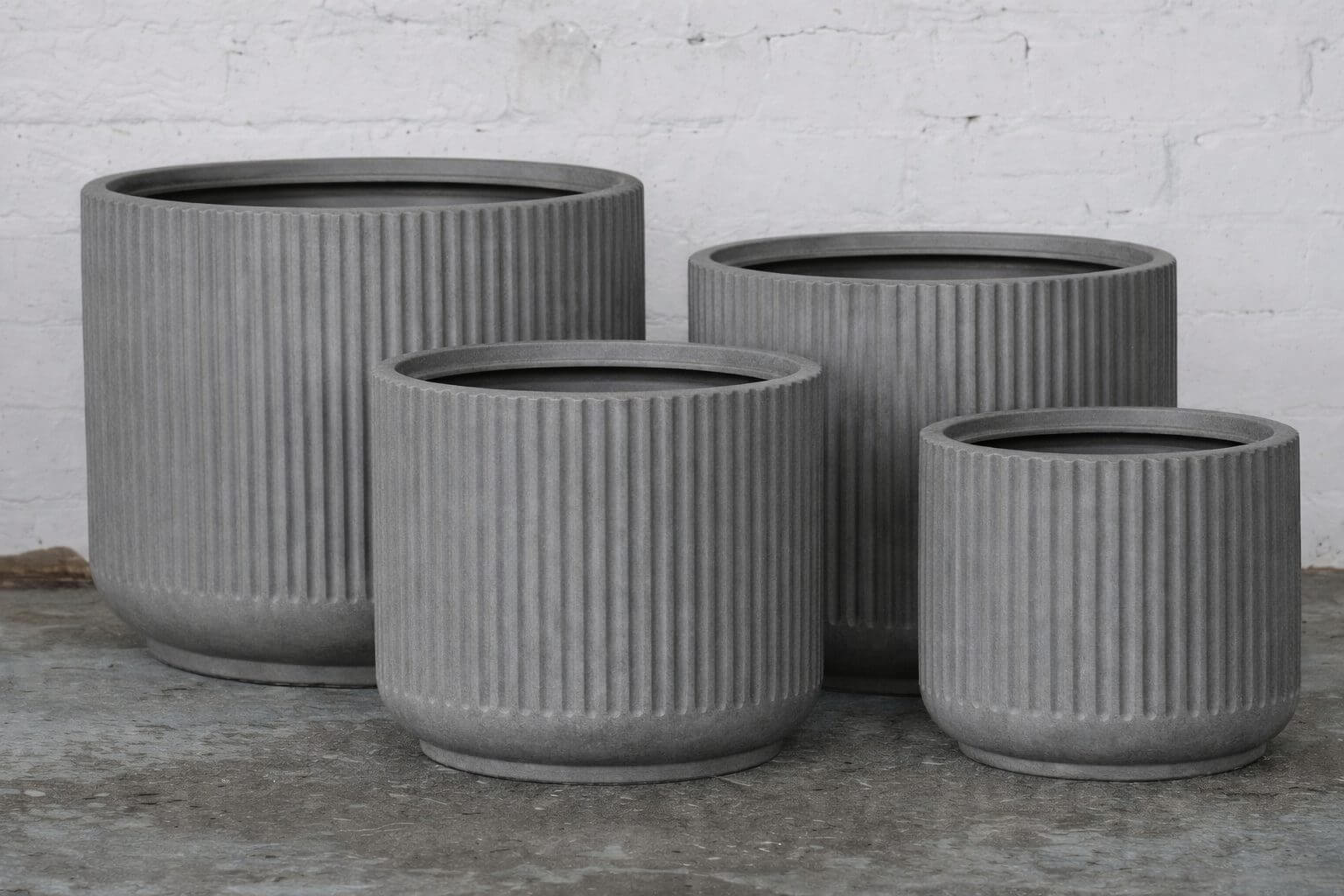 Four grey, ribbed, cylindrical planters of varying sizes arranged on a concrete floor against a white brick wall.