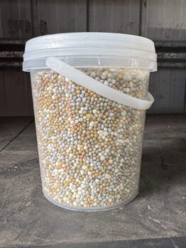 A clear bucket of Fertiliser Slow Release 1kg, filled with multicoloured granular pellets, sits on a dusty surface in front of a corrugated metal wall.