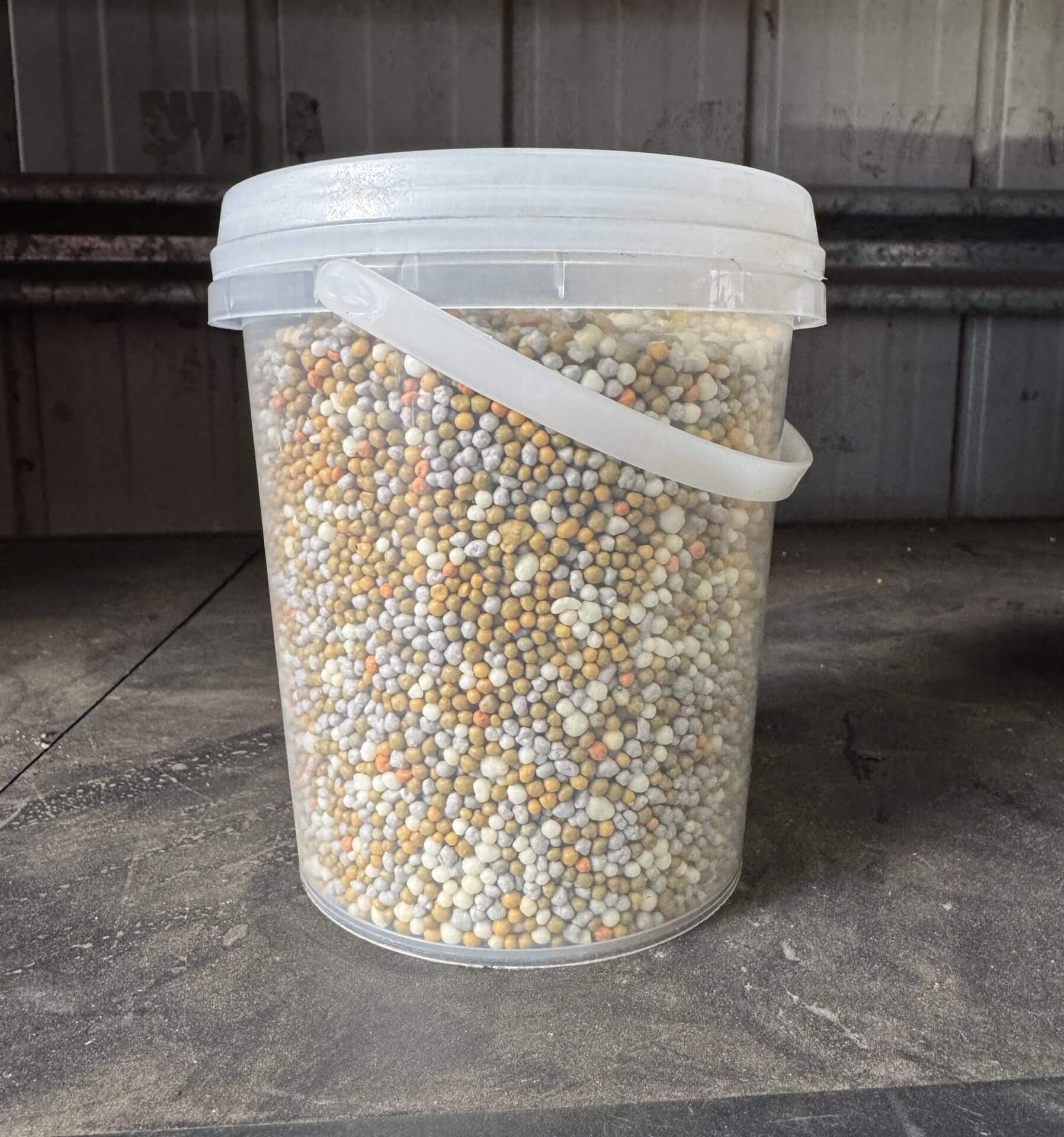 A clear bucket of Fertiliser Slow Release 1kg, filled with multicoloured granular pellets, sits on a dusty surface in front of a corrugated metal wall.