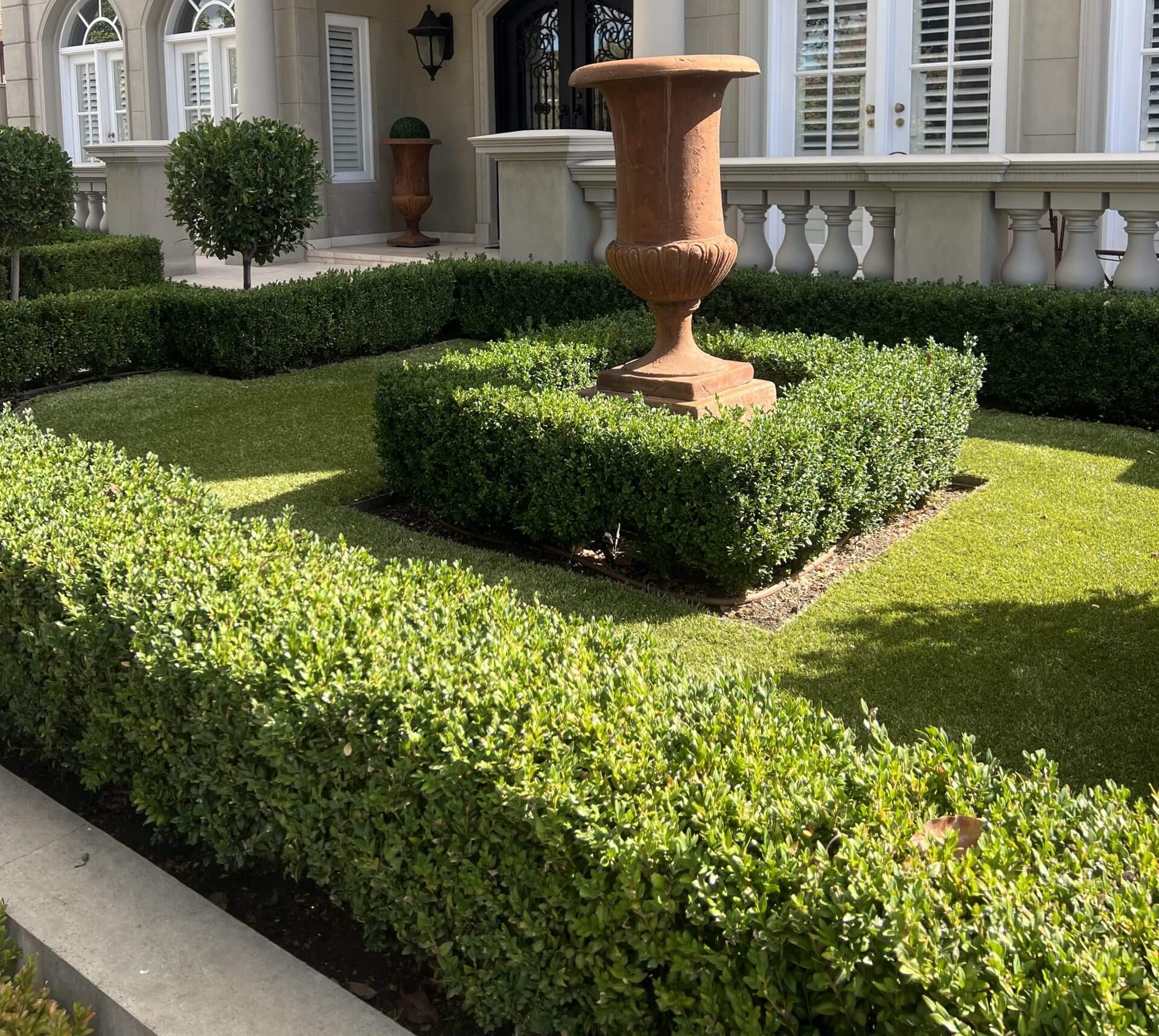 A formal garden with neatly trimmed hedges, a square lawn area, and a large decorative urn on a plinth in front of an elegant building.